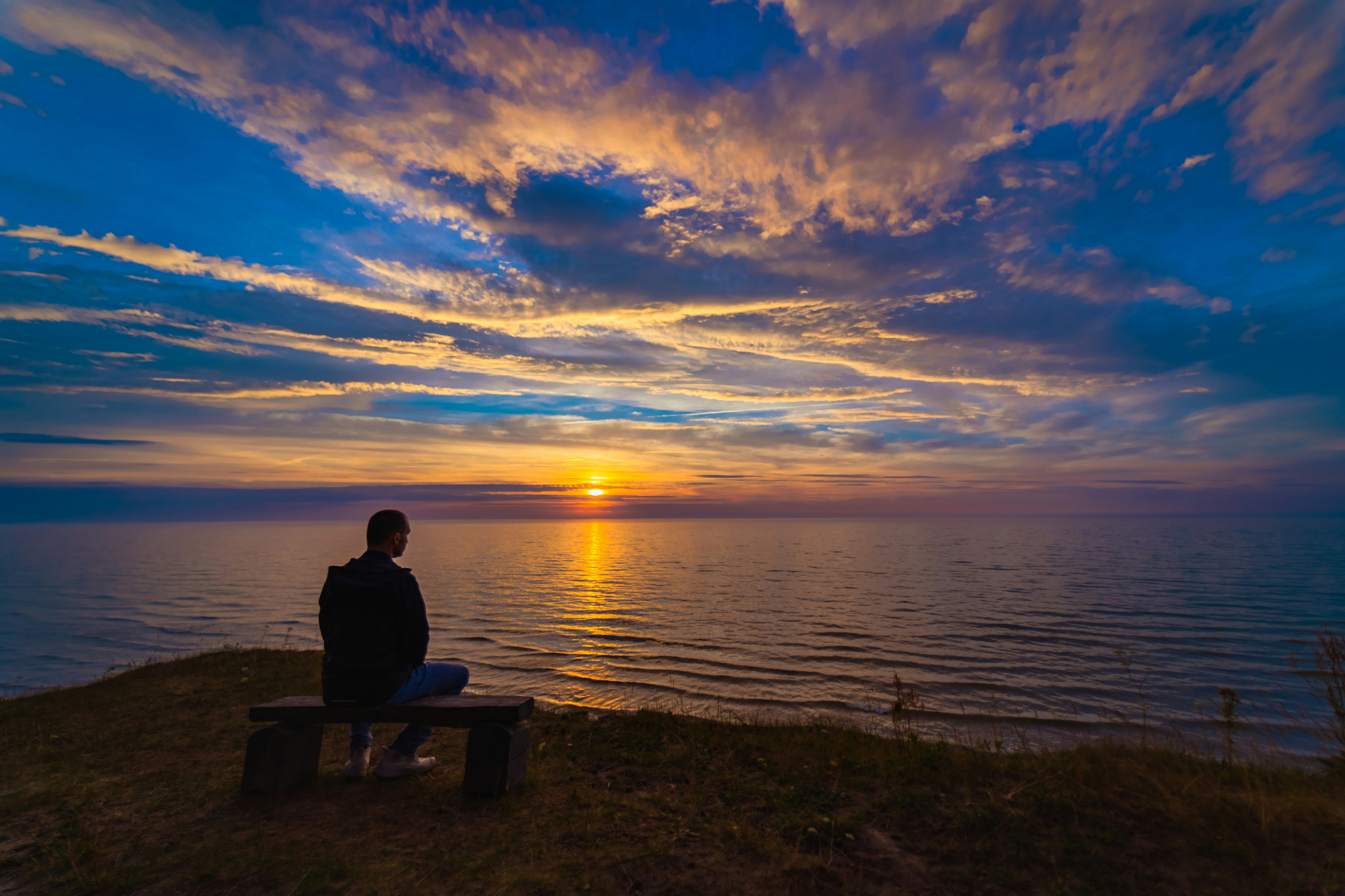 man sitting on bench near body of water during sunset