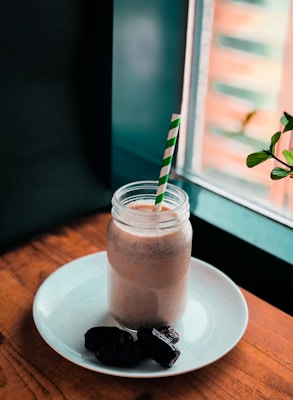 A creamy beverage in a mason jar with a green and white striped straw, placed on a white plate with three dates. The setup is on a wooden surface near a window, with some green leaves visible in the background.