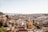 A panoramic view of a cityscape featuring a mix of historic and modern buildings with red-tiled roofs. In the foreground, there are densely packed buildings, some with colorful facades. A prominent yellow building stands out. In the background, a suspension bridge similar to the Golden Gate Bridge spans across a body of water leading out to the horizon.