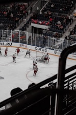 A hockey game is taking place in an indoor stadium. Players in red and white uniforms are actively engaged near the goal, with one team seemingly attempting to score. The stands are partially filled with spectators, and there is visible glass shielding surrounding the rink.