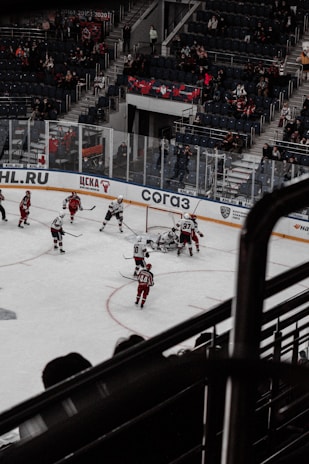 A hockey game is taking place in an indoor stadium. Players in red and white uniforms are actively engaged near the goal, with one team seemingly attempting to score. The stands are partially filled with spectators, and there is visible glass shielding surrounding the rink.