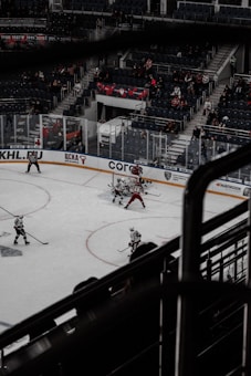 A hockey game is taking place in an indoor arena with players wearing uniforms in white and red. The action is centered near the goal as players compete for the puck. Spectators are visible in the stands, although the arena is not fully packed.