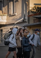 A group of young people walks together on a street next to a building. They are carrying backpacks and wearing casual clothes. The mood appears friendly as one person gestures with a peace sign while others are engaged with each other.