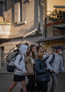 A group of young people walks together on a street next to a building. They are carrying backpacks and wearing casual clothes. The mood appears friendly as one person gestures with a peace sign while others are engaged with each other.