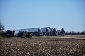 A rural landscape featuring a large agricultural building with four metallic silos. In the foreground, a green tractor is connected to farming equipment, preparing a field. The background includes flat, open land with sparse trees and a clear blue sky.