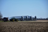 A rural landscape featuring a large agricultural building with four metallic silos. In the foreground, a green tractor is connected to farming equipment, preparing a field. The background includes flat, open land with sparse trees and a clear blue sky.