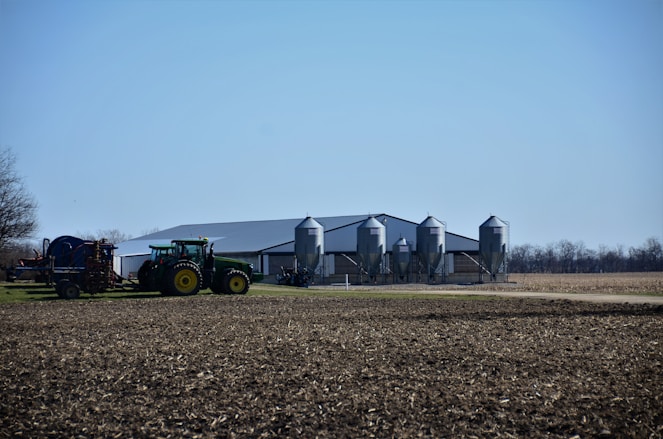 A rural landscape featuring a large agricultural building with four metallic silos. In the foreground, a green tractor is connected to farming equipment, preparing a field. The background includes flat, open land with sparse trees and a clear blue sky.