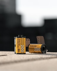 Two rolls of Kodak film are positioned on a surface. The foreground film roll displays the details 'Kodacolor 200', and '35mm Film for Color Prints'. Another film roll in the background is slightly out of focus. Both rolls have a distinct yellow and black design, characteristic of Kodak film packaging. The setting is out of focus, suggesting an indoor environment with natural lighting.