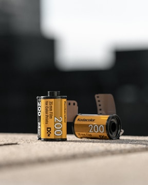 Two rolls of Kodak film are positioned on a surface. The foreground film roll displays the details 'Kodacolor 200', and '35mm Film for Color Prints'. Another film roll in the background is slightly out of focus. Both rolls have a distinct yellow and black design, characteristic of Kodak film packaging. The setting is out of focus, suggesting an indoor environment with natural lighting.