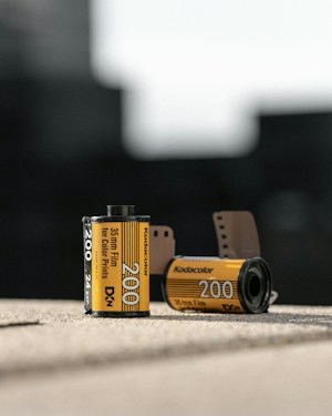 Two rolls of Kodak film are positioned on a surface. The foreground film roll displays the details 'Kodacolor 200', and '35mm Film for Color Prints'. Another film roll in the background is slightly out of focus. Both rolls have a distinct yellow and black design, characteristic of Kodak film packaging. The setting is out of focus, suggesting an indoor environment with natural lighting.