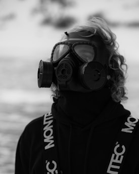 A person wearing a gas mask stands in an outdoor setting. The mask features large circular filters on both sides and covers the entire face, while the individual wears a dark sweater with the word 'MONTEC' visible on the shoulder straps. The background appears to be blurred, possibly indicating snow or a neutral setting.