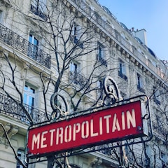 An ornate Metropolitain sign in front of a classic Haussmann-style building with intricate balconies and bare tree branches reaching up towards a clear blue sky.