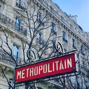 An ornate Metropolitain sign in front of a classic Haussmann-style building with intricate balconies and bare tree branches reaching up towards a clear blue sky.