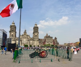 A large, historic cathedral with two towers stands prominently against a partly cloudy sky. In the foreground, a Mexican flag is flying, and a cannon is placed behind a series of green metal barriers. Many people are walking around the open plaza, some taking photographs or gathering in groups.