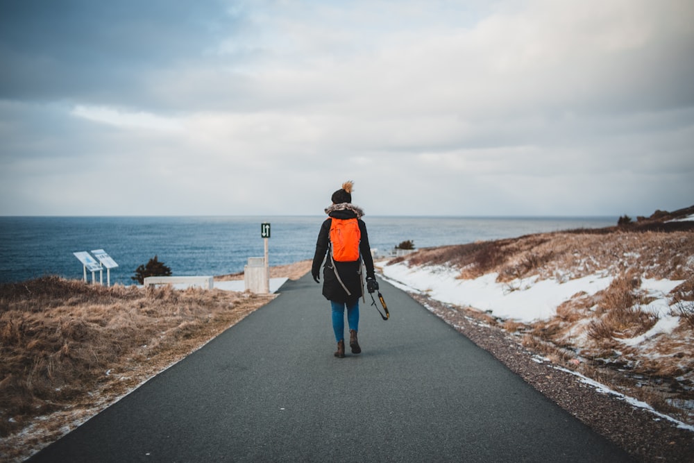 woman in black jacket and blue denim jeans walking on gray concrete pathway near sea during