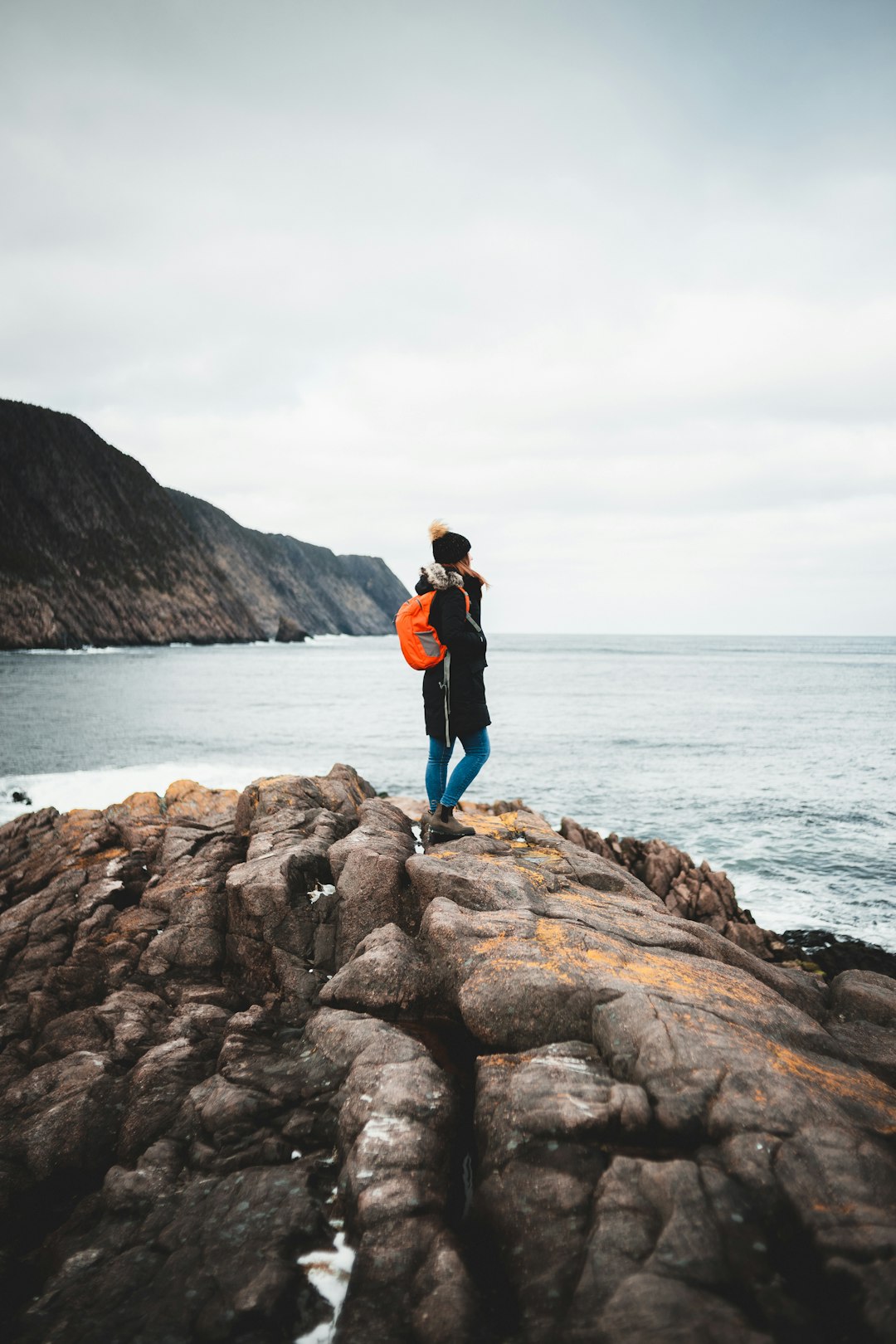 woman in red jacket and blue denim jeans standing on brown rock near body of water