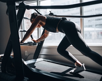 A person with a tattooed arm is engaged in a workout on a treadmill, using their hands for support. The room is brightly lit by natural light from the large window, highlighting the athletic wear and muscular build of the individual.