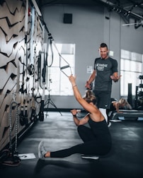 A woman in athletic wear is sitting on the floor of a gym, engaging with suspension training ropes attached to the wall. She appears to be stretching or exercising, while a man in a t-shirt is standing nearby, possibly offering guidance or instructions. The gym setting includes exercise equipment and natural light coming through large windows.
