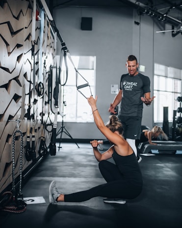 A trainer guiding a young man through a stretching routine in a bright, modern gym space.