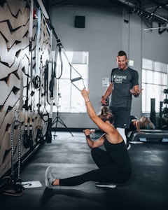 A woman in athletic wear is sitting on the floor of a gym, engaging with suspension training ropes attached to the wall. She appears to be stretching or exercising, while a man in a t-shirt is standing nearby, possibly offering guidance or instructions. The gym setting includes exercise equipment and natural light coming through large windows.