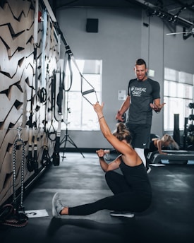 A woman in athletic wear is sitting on the floor of a gym, engaging with suspension training ropes attached to the wall. She appears to be stretching or exercising, while a man in a t-shirt is standing nearby, possibly offering guidance or instructions. The gym setting includes exercise equipment and natural light coming through large windows.
