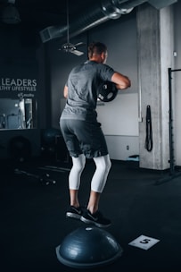 A person is performing a balance exercise in a gym setting. They are dressed in athletic wear, standing on a Bosu ball while holding a weight plate. The environment includes gym equipment and motivational wall text.