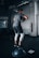 A person is performing a balance exercise in a gym setting. They are dressed in athletic wear, standing on a Bosu ball while holding a weight plate. The environment includes gym equipment and motivational wall text.