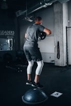 A person is performing a balance exercise in a gym setting. They are dressed in athletic wear, standing on a Bosu ball while holding a weight plate. The environment includes gym equipment and motivational wall text.