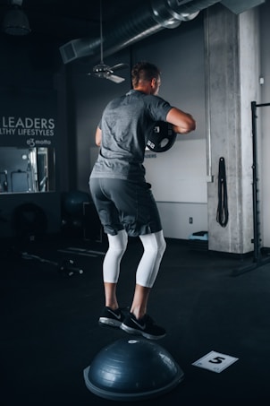 A person is performing a balance exercise in a gym setting. They are dressed in athletic wear, standing on a Bosu ball while holding a weight plate. The environment includes gym equipment and motivational wall text.