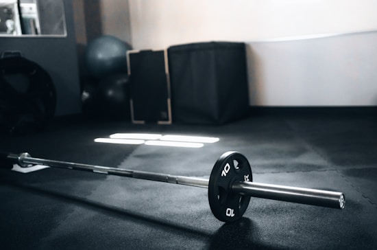 A barbell with weight plates is resting on a dark rubber gym floor. In the background, there are exercise balls and fitness equipment such as padding and mats, lit by natural light coming through a window, creating a soft reflection on the floor.