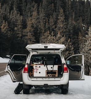 An SUV is parked on a snowy path with its rear doors open, revealing camping or travel gear neatly organized inside. The scene is set against a backdrop of dense evergreen trees, suggesting a forest location.