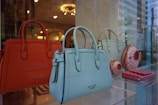 A display of luxury handbags is showcased in a window, featuring vibrant orange, blue, and green tones. The bags have elegant designs, with some resembling confectionery items. The background reflects the interior of a well-lit store with decorative lighting.