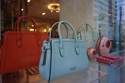 A display of luxury handbags is showcased in a window, featuring vibrant orange, blue, and green tones. The bags have elegant designs, with some resembling confectionery items. The background reflects the interior of a well-lit store with decorative lighting.