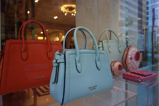 A display of luxury handbags is showcased in a window, featuring vibrant orange, blue, and green tones. The bags have elegant designs, with some resembling confectionery items. The background reflects the interior of a well-lit store with decorative lighting.