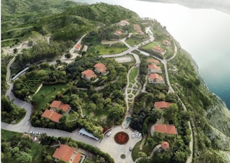 An aerial view of a landscaped area featuring several houses with red roofs surrounded by lush greenery and winding roads. The setting appears to be on a hillside by a large body of water, with well-maintained gardens and pathways. The terrain is hilly, with dense forestation in some areas and open grass fields in others. Cars can be seen parked around the circular driveway at the center of the scene.