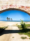A stone archway with red graffiti reading 'DIAS BONS VIRÃO' frames a view of a beach. Beyond the arch, a concrete path leads toward a sandy area with a few palm trees. The sky is clear and blue, with the ocean visible in the distance. Green foliage grows at the sides of the path, adding a touch of nature to the scene.