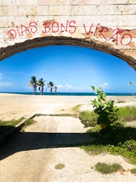 A stone archway with red graffiti reading 'DIAS BONS VIRÃO' frames a view of a beach. Beyond the arch, a concrete path leads toward a sandy area with a few palm trees. The sky is clear and blue, with the ocean visible in the distance. Green foliage grows at the sides of the path, adding a touch of nature to the scene.