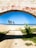 A stone archway with red graffiti reading 'DIAS BONS VIRÃO' frames a view of a beach. Beyond the arch, a concrete path leads toward a sandy area with a few palm trees. The sky is clear and blue, with the ocean visible in the distance. Green foliage grows at the sides of the path, adding a touch of nature to the scene.