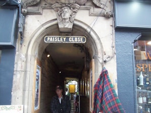 An arched stone entrance sign reading 'Paisley Close'. A decorative stone carving of a person is above the arch. Below the arch, a person is partially in shadow. A window display with wine bottles and a plaid cloth is visible nearby.
