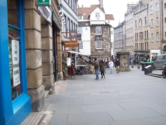 A street scene featuring historical architecture with several buildings, including a pharmacy and a café named Monteith's. People are walking and standing on the cobbled street. Visible in the foreground is a blue storefront with a no-smoking sign that also offers a free stop smoking service.