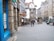 A street scene featuring historical architecture with several buildings, including a pharmacy and a café named Monteith's. People are walking and standing on the cobbled street. Visible in the foreground is a blue storefront with a no-smoking sign that also offers a free stop smoking service.