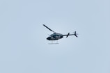 A helicopter is flying in the sky, set against a pale blue background. The helicopter appears to be dark in color and is captured in mid-air, showing details of its rotor blades and tail.
