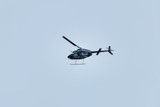 A helicopter is flying in the sky, set against a pale blue background. The helicopter appears to be dark in color and is captured in mid-air, showing details of its rotor blades and tail.