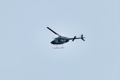 A helicopter is flying in the sky, set against a pale blue background. The helicopter appears to be dark in color and is captured in mid-air, showing details of its rotor blades and tail.