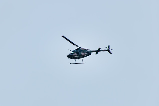 A helicopter is flying in the sky, set against a pale blue background. The helicopter appears to be dark in color and is captured in mid-air, showing details of its rotor blades and tail.
