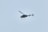 A helicopter is flying in the sky, set against a pale blue background. The helicopter appears to be dark in color and is captured in mid-air, showing details of its rotor blades and tail.