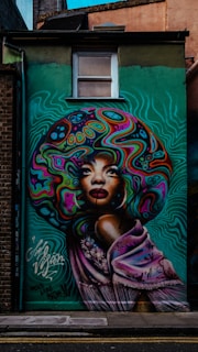 A vibrant mural featuring a stylized portrait of a woman with an elaborate, colorful afro. The design incorporates swirling patterns and bold colors in the hair, with a serene expression on her face. Her shoulders are draped in a lilac shawl decorated with floral motifs. The background is a gradient of teal green with wavy lines, enhancing the dynamic feel.