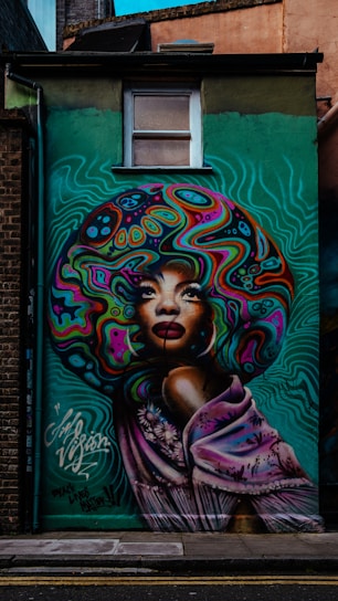 A vibrant mural featuring a stylized portrait of a woman with an elaborate, colorful afro. The design incorporates swirling patterns and bold colors in the hair, with a serene expression on her face. Her shoulders are draped in a lilac shawl decorated with floral motifs. The background is a gradient of teal green with wavy lines, enhancing the dynamic feel.