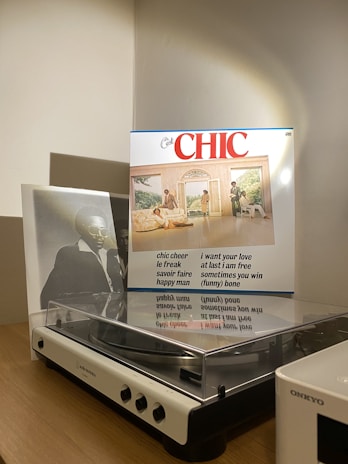 A vintage turntable sits on a wooden surface, next to a vinyl record cover featuring a group of people in a stylish, retro setting. The cover includes a list of song titles and the word 'CHIC' prominently displayed at the top. The lighting casts a warm glow, highlighting the turntable and the record.