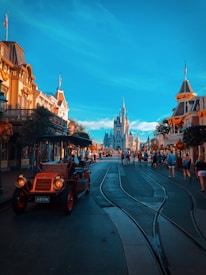 A vibrant street scene in a theme park, featuring a vintage car in the foreground on tram tracks. The street is lined with buildings adorned with festive decorations, and a castle stands prominently in the background. Visitors are leisurely walking along the street, enjoying the bright, clear day.
